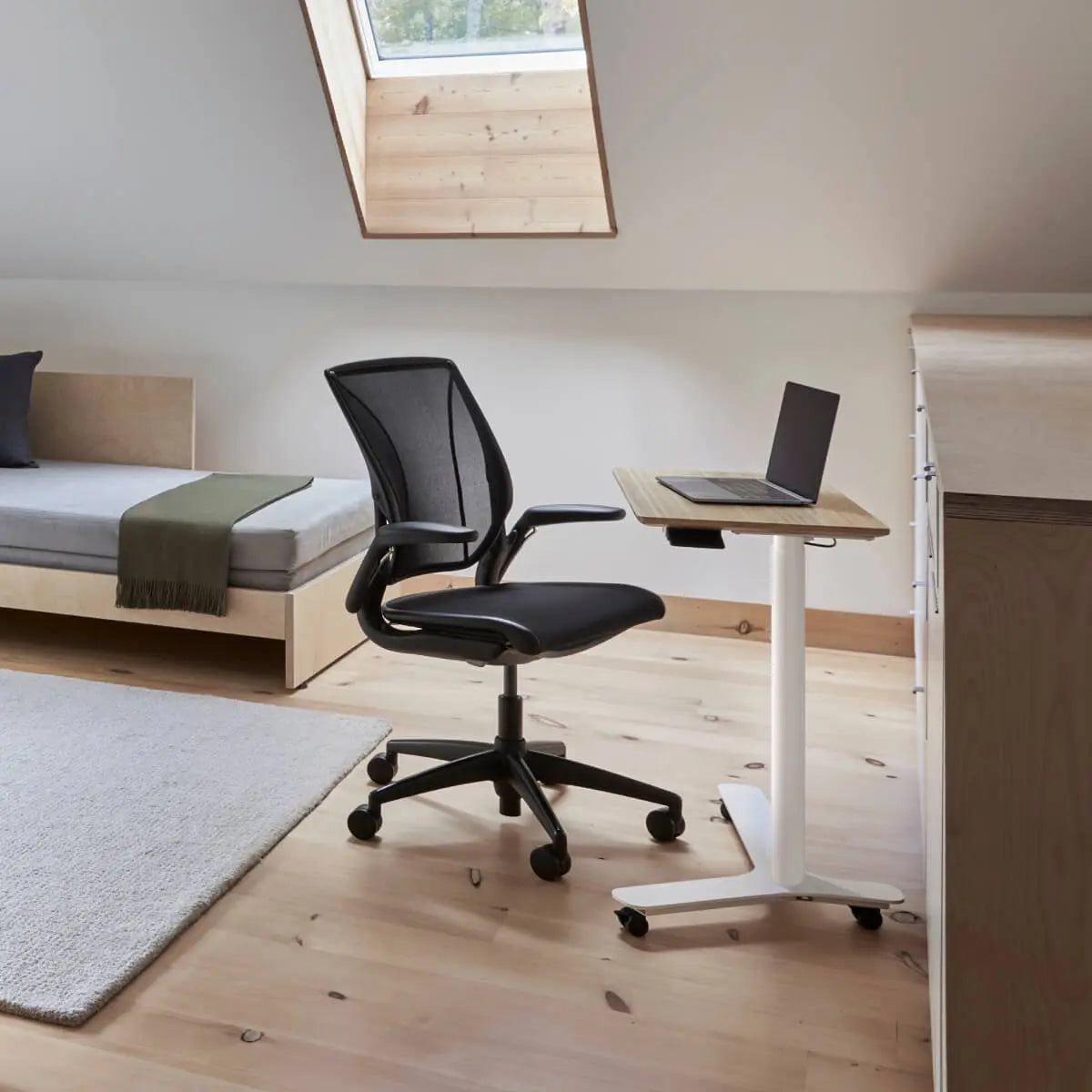 Modern office chair and adjustable desk in a room with a bed and window.