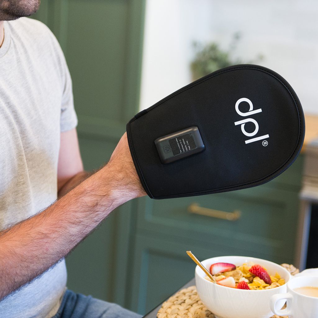 A person in a light gray t-shirt holds a black red light therapy hand mitt with the white 'dpl' logo, set against a blurred kitchen background with a bowl of cereal on the table.