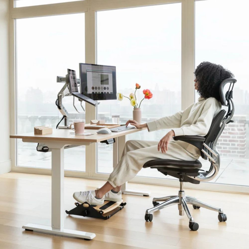 Woman seated in an ergonomic office chair at a standing desk with dual monitors in a bright modern workspace.