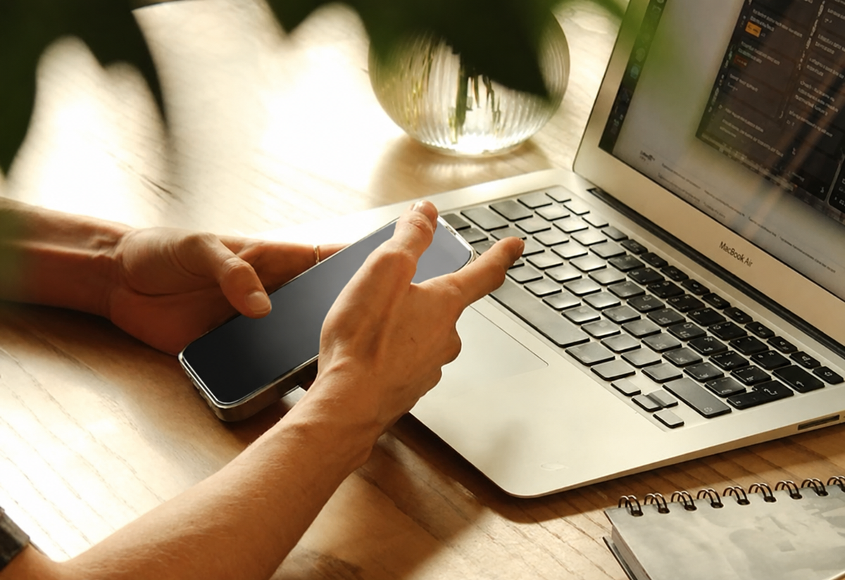 Person using a smartphone beside an open laptop on a sunlit wooden desk.