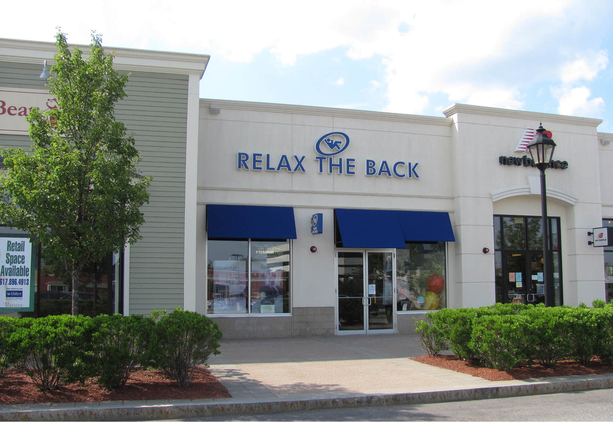 Relax The Back retail storefront with blue awnings, glass entrance, and the store name in blue lettering on a white facade, photographed outdoors with trees and shrubs in front.
