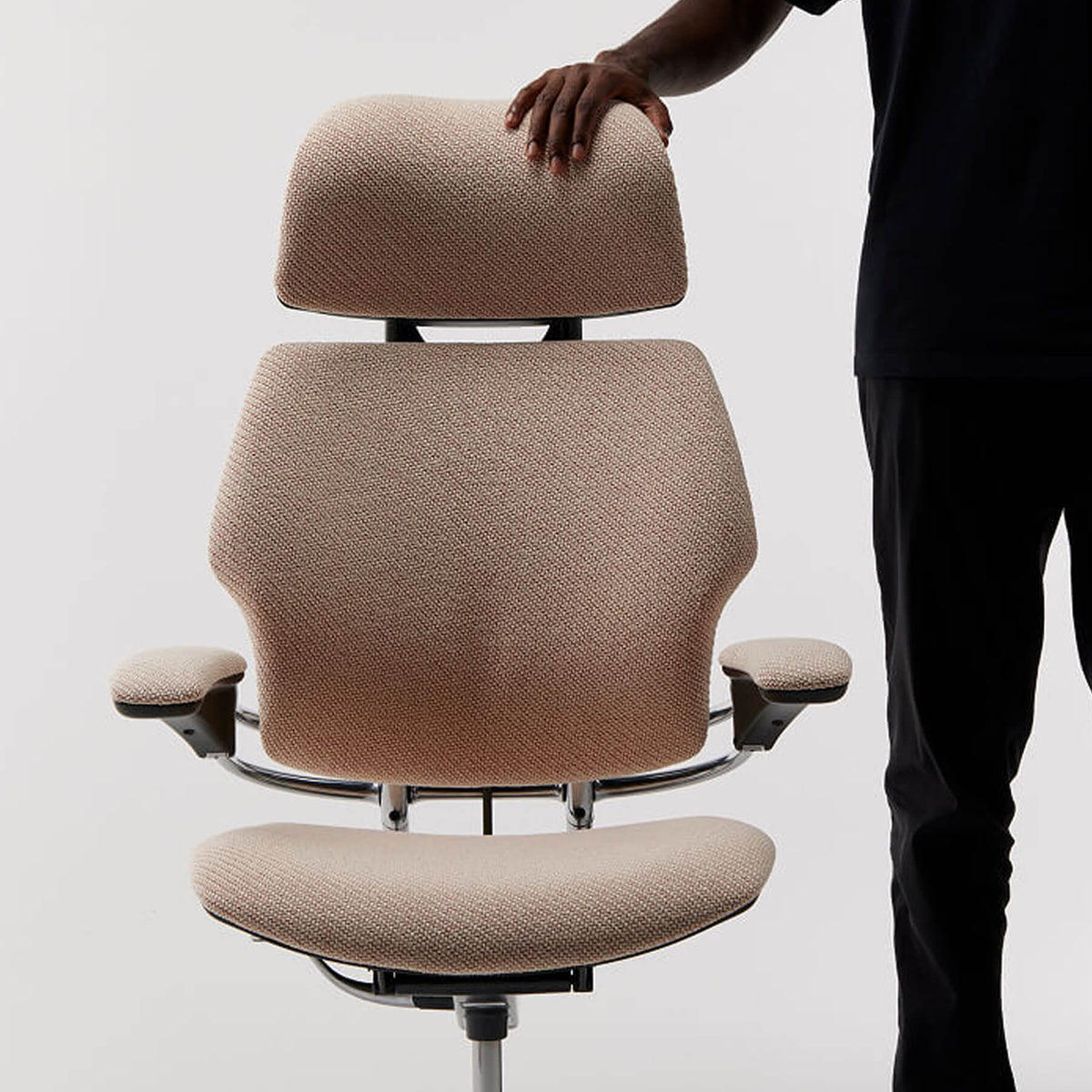 A beige upholstered ergonomic office chair with a headrest, armrests, and a curved back, shown against a light gray studio background. A person's hand and legs are visible on the right, touching the headrest.