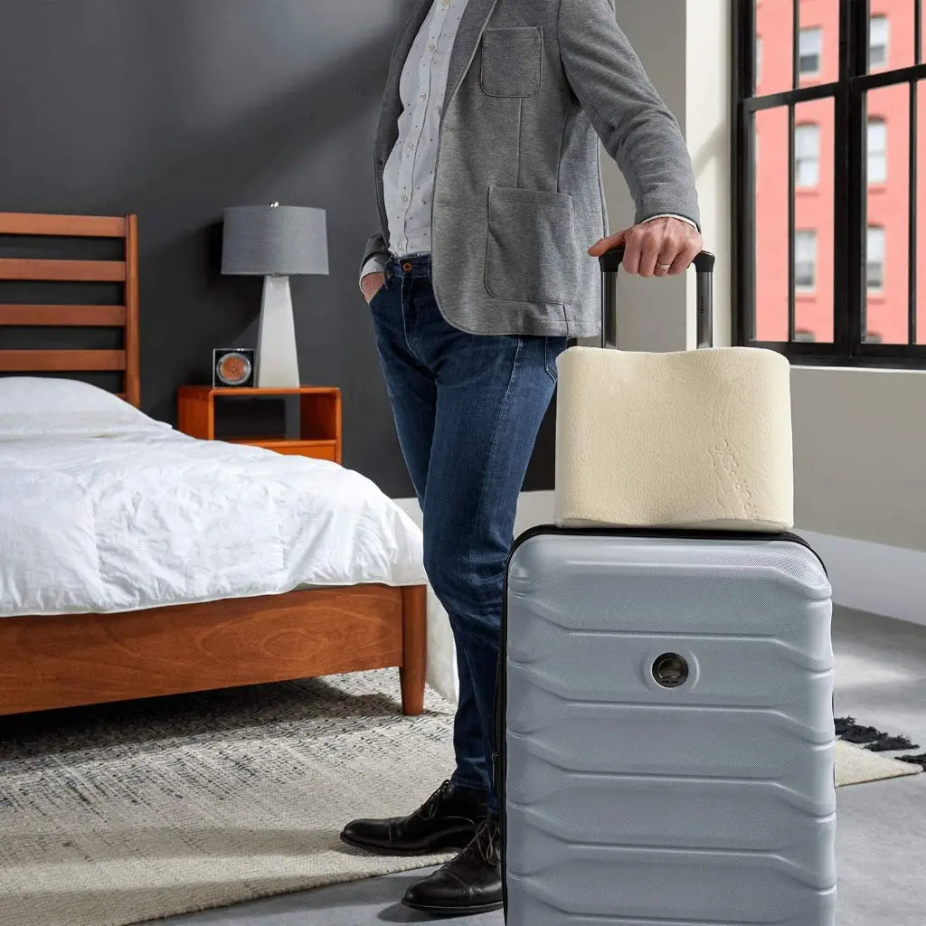 A man standing in a bedroom holding his suit case and the Tempur-Pedic Travel Neck Pillow.