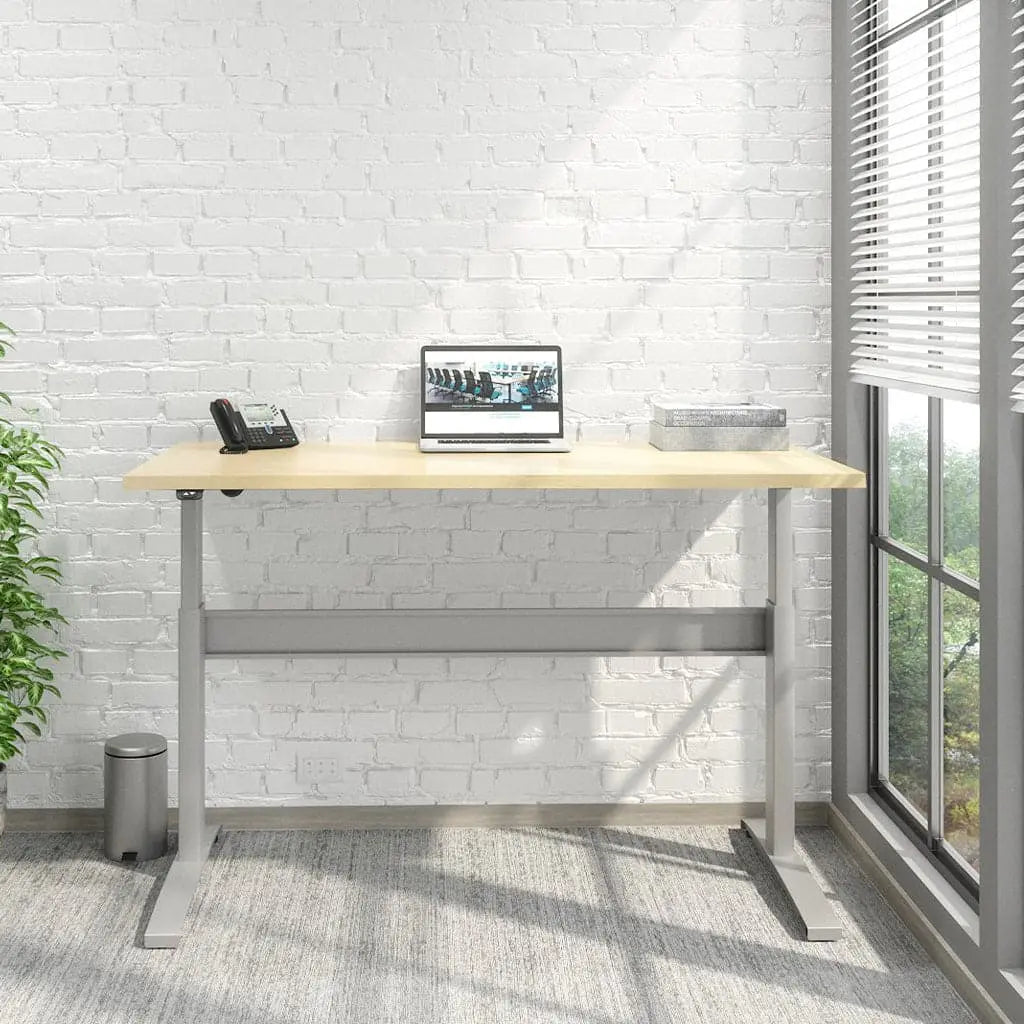 A Transcend Desk positioned beside a window features a laptop and phone on its surface. The desk boasts a wooden top paired with a sleek silver base.