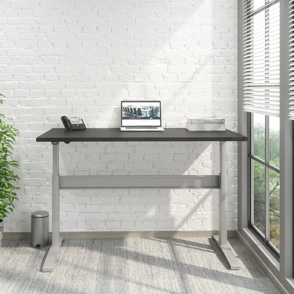A Transcend Desk positioned beside a window features a laptop and phone on its surface. The desk boasts a black wooden top paired with a sleek silver base.