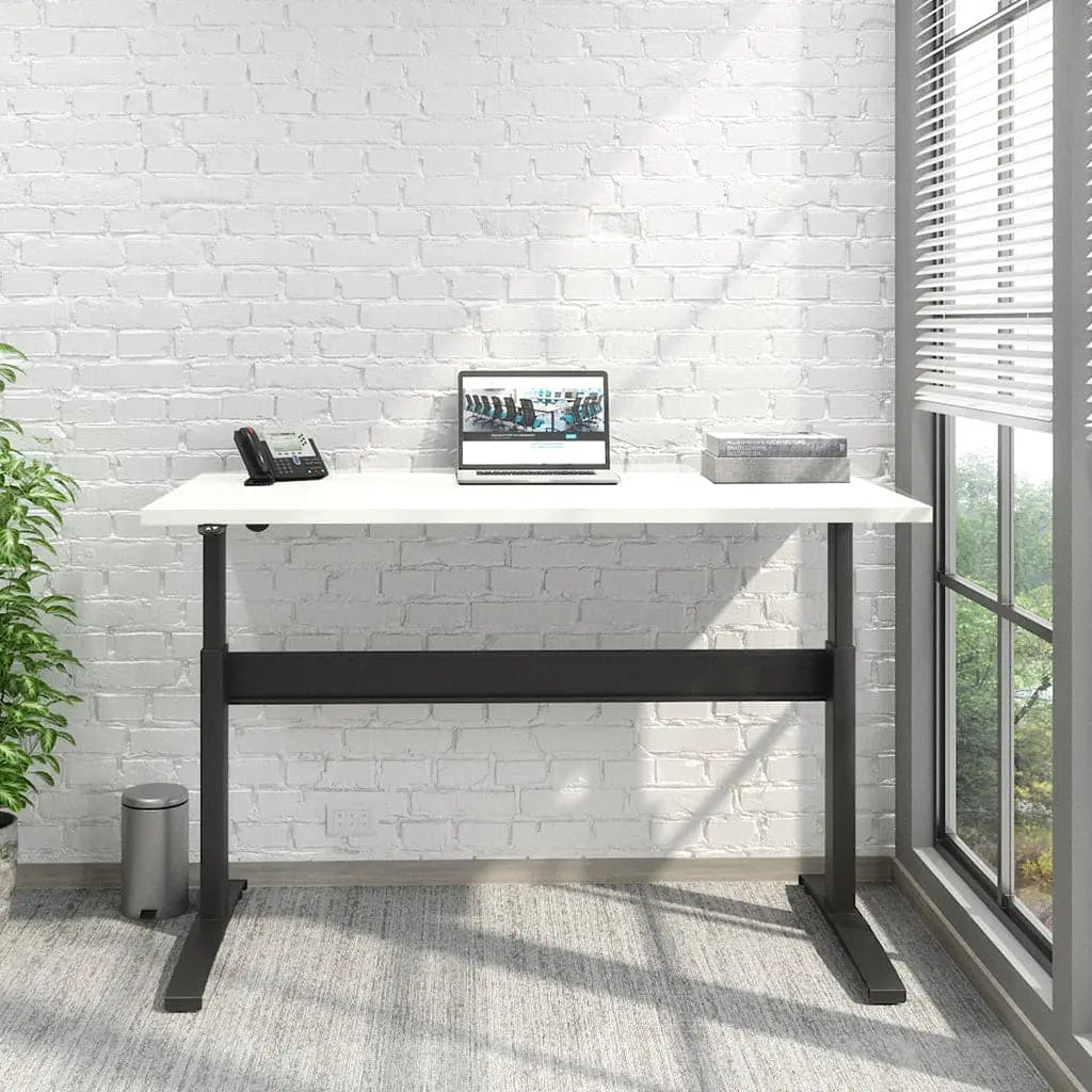 A Transcend Desk positioned beside a window features a laptop and phone on its surface. The desk boasts a white wooden top paired with a sleek black base.