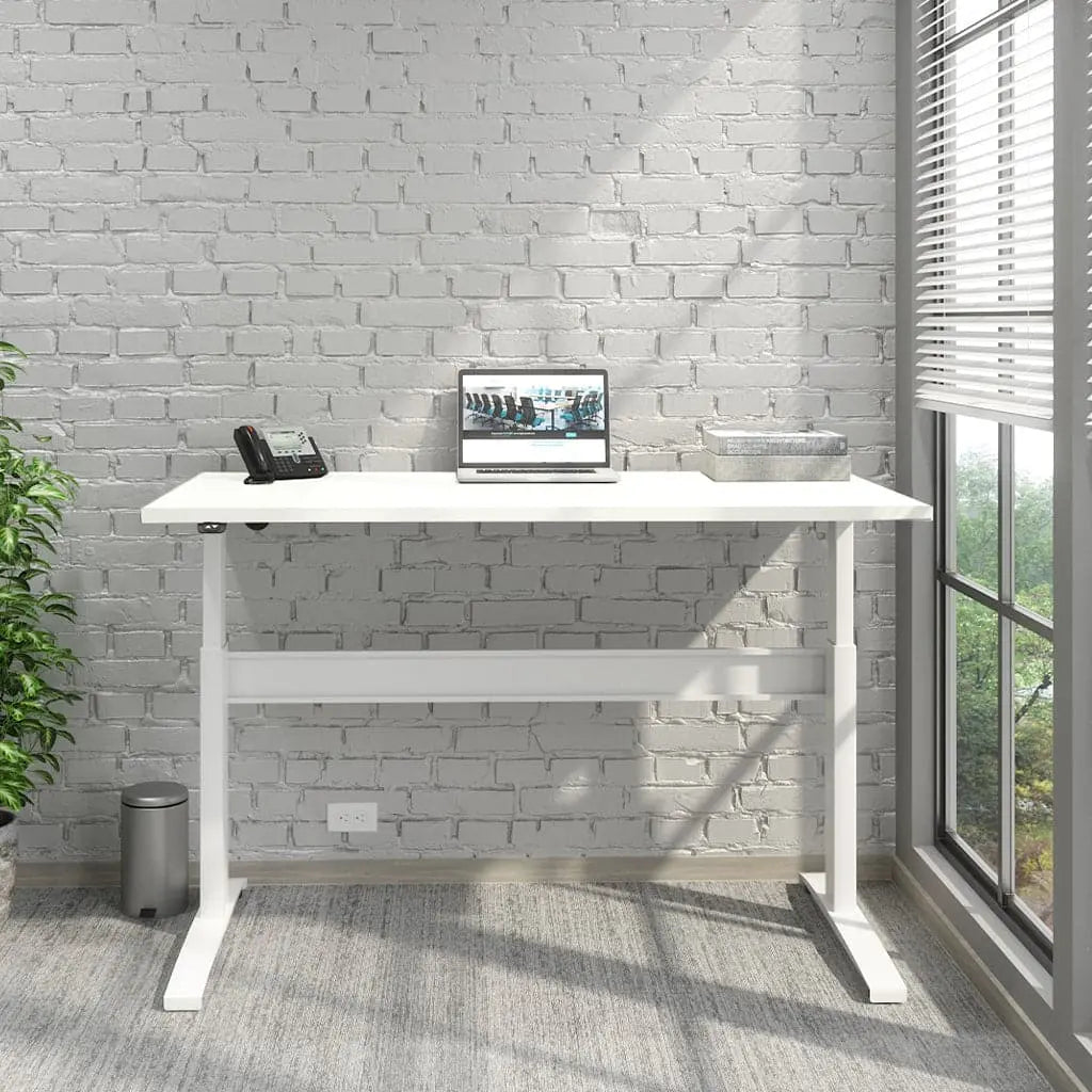 A Transcend Desk positioned beside a window features a laptop and phone on its surface. The desk boasts a white wooden top paired with a sleek white base.