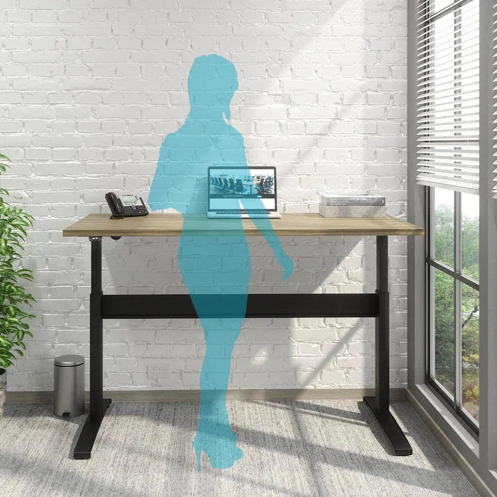 A modern Transcend desk, featuring a wood top and a black metal frame, is situated in a bright office with a white brick wall and a large window with blinds. On the desk are a telephone, a laptop displaying an office scene, and a box or organizer. A translucent blue silhouette of a person stands at the desk, suggesting that the desk is used in a standing position. A small trash can and a plant are visible in the background, and sunlight streams into the room.